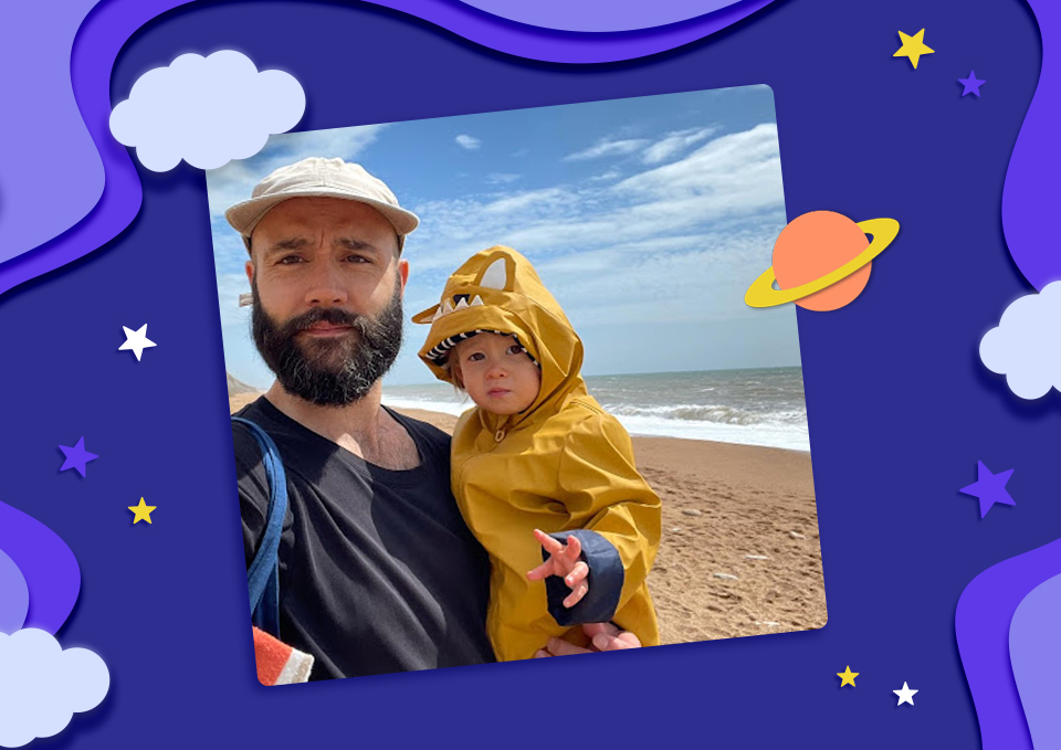 Father holding his toddler daughter on a sandy beach - she is wearing a yellow raincoat. The picture's backdrop is blue illustrated space and planets